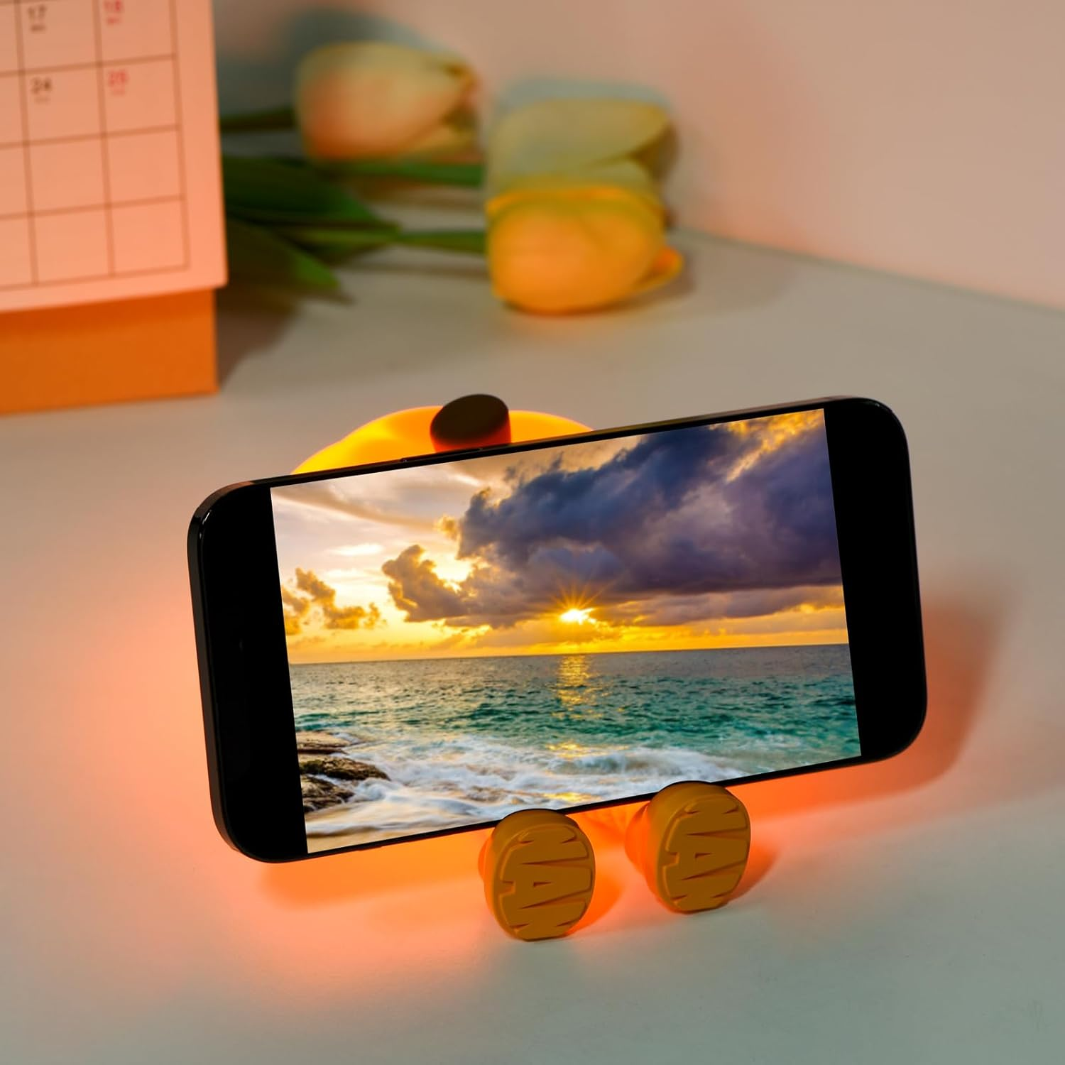 Un telefono che mostra un tramonto sull'oceano, su un supporto giallo. Un calendario e un tulipano sullo sfondo.