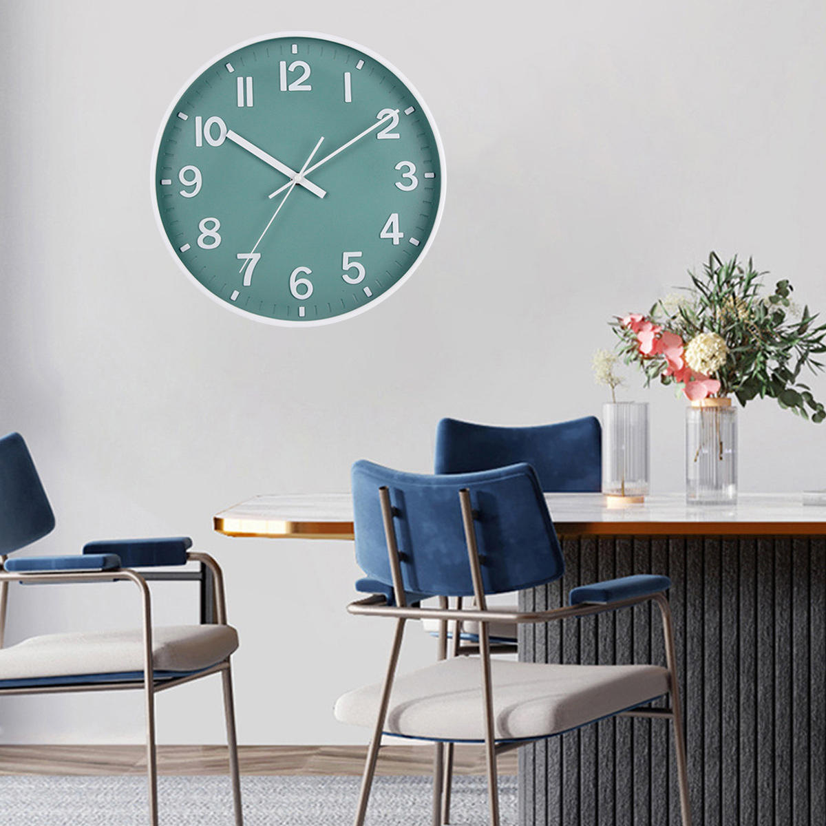 Un orologio turchese è appeso a un muro bianco. Sotto, un tavolo con sedie e un vaso di fiori.