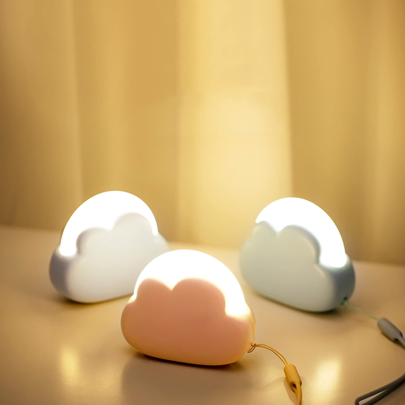 Tre luci notturne a forma di nuvola. Una è rosa, le altre bianche e blu. Brillano su un tavolo.