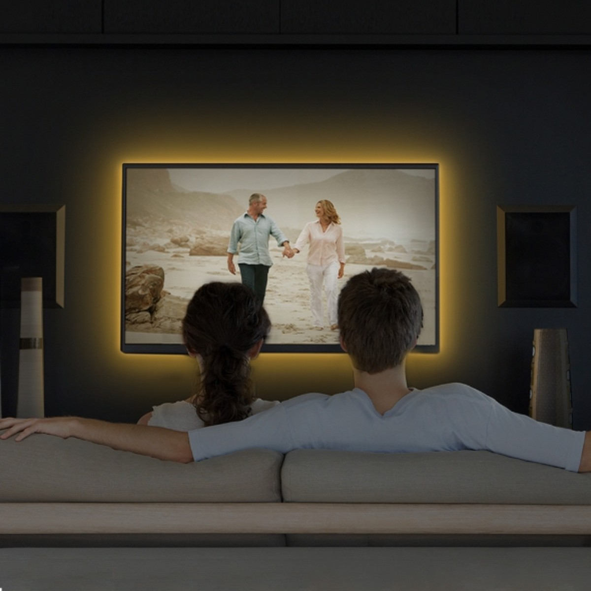 Para ogląda telewizję. Na ekranie para spaceruje po plaży. Telewizor otoczony jest pomarańczową poświatą. Siedzą na kanapie.