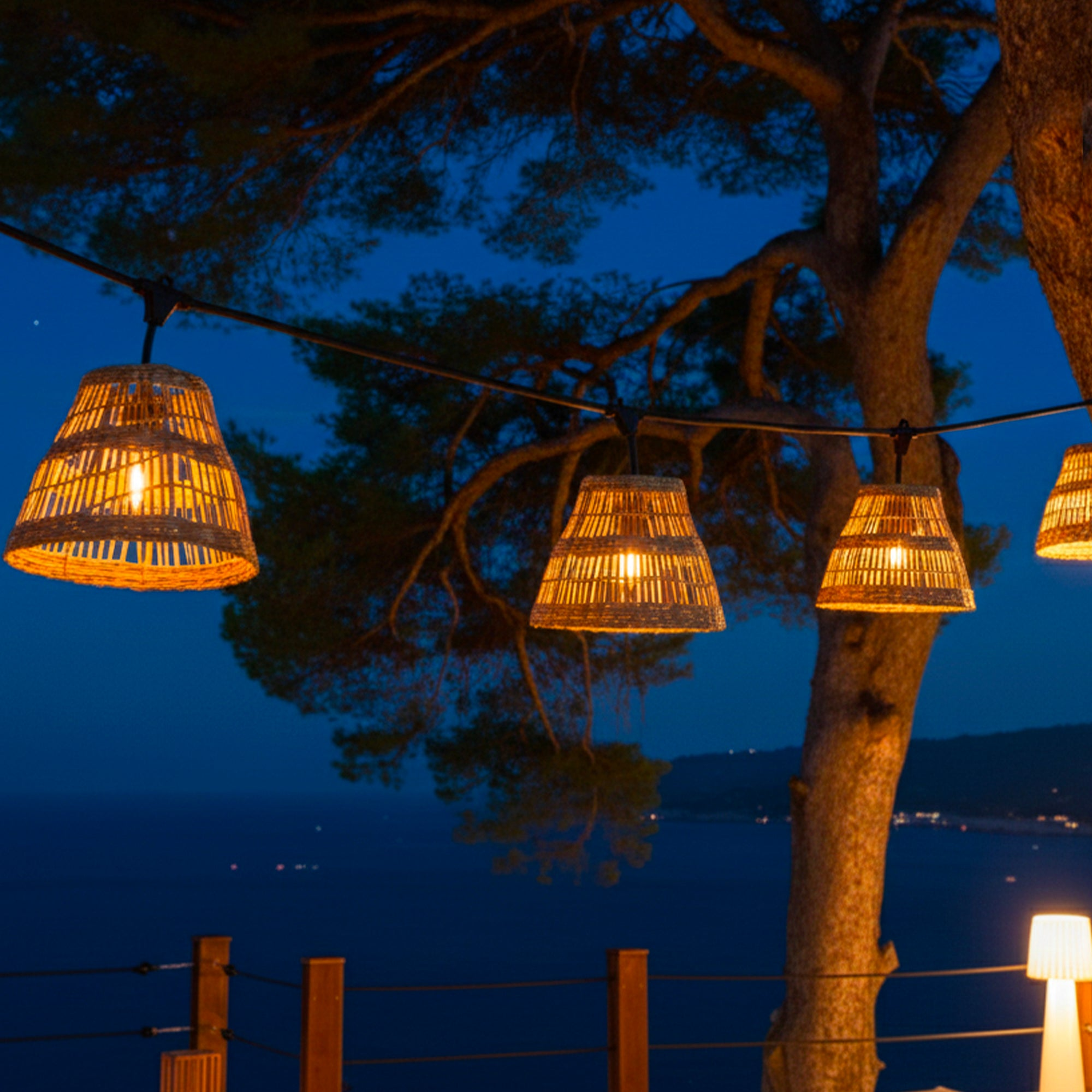 Abendliche Szene mit Lichterketten, Baum und Meer im Hintergrund. Die Lichter sind Korblampenschirme, die gegen den dunkelblauen Himmel leuchten.