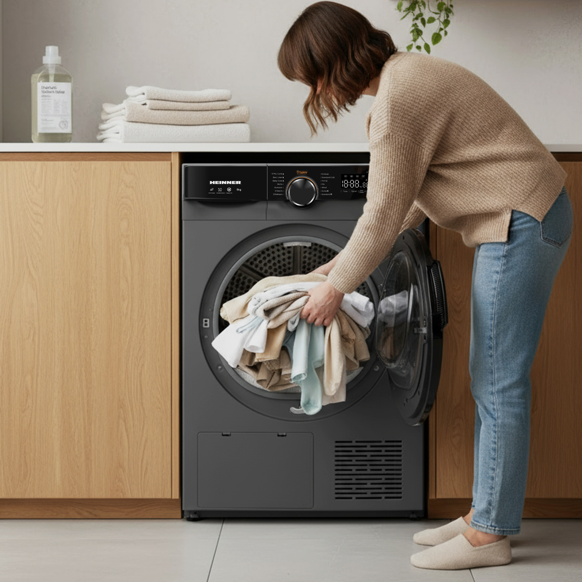 A woman loads a gray dryer with clothes. She wears jeans and a sweater. A bottle and towels are on a counter.