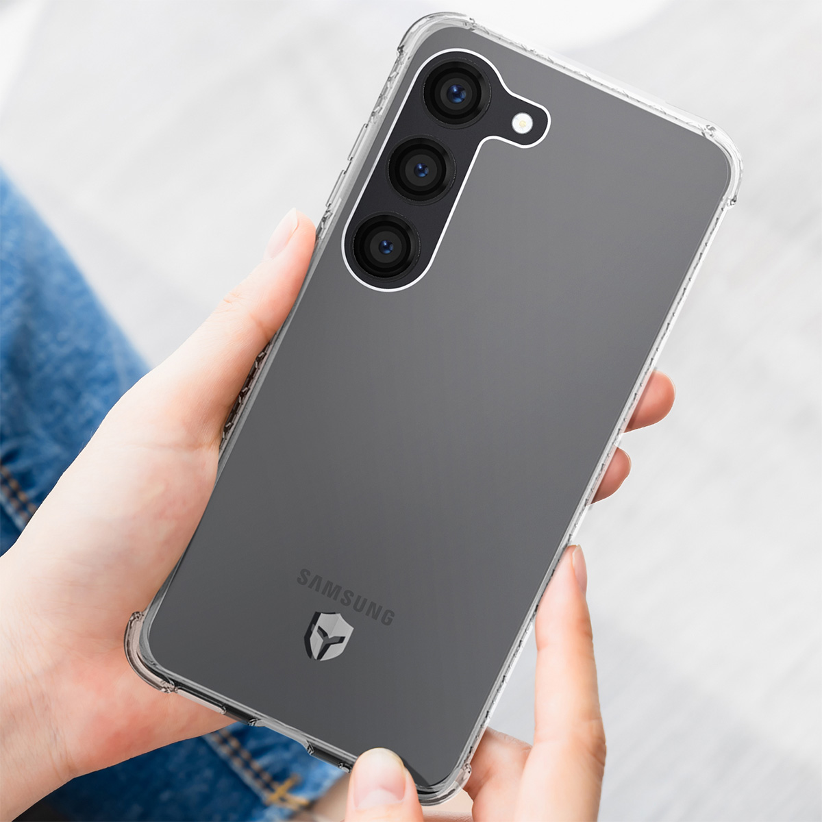 Een persoon houdt een doorzichtige telefoonhoes vast. De hoes omsluit een donkergrijze telefoon met een drievoudige cameralens en het Samsung-logo.