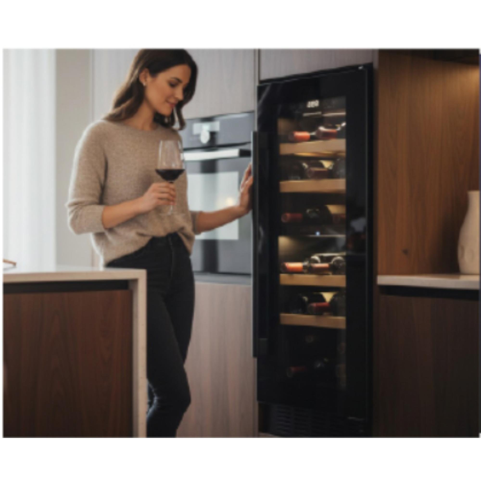 A woman holds a wine glass, looking at a wine fridge filled with bottles, in a kitchen setting.