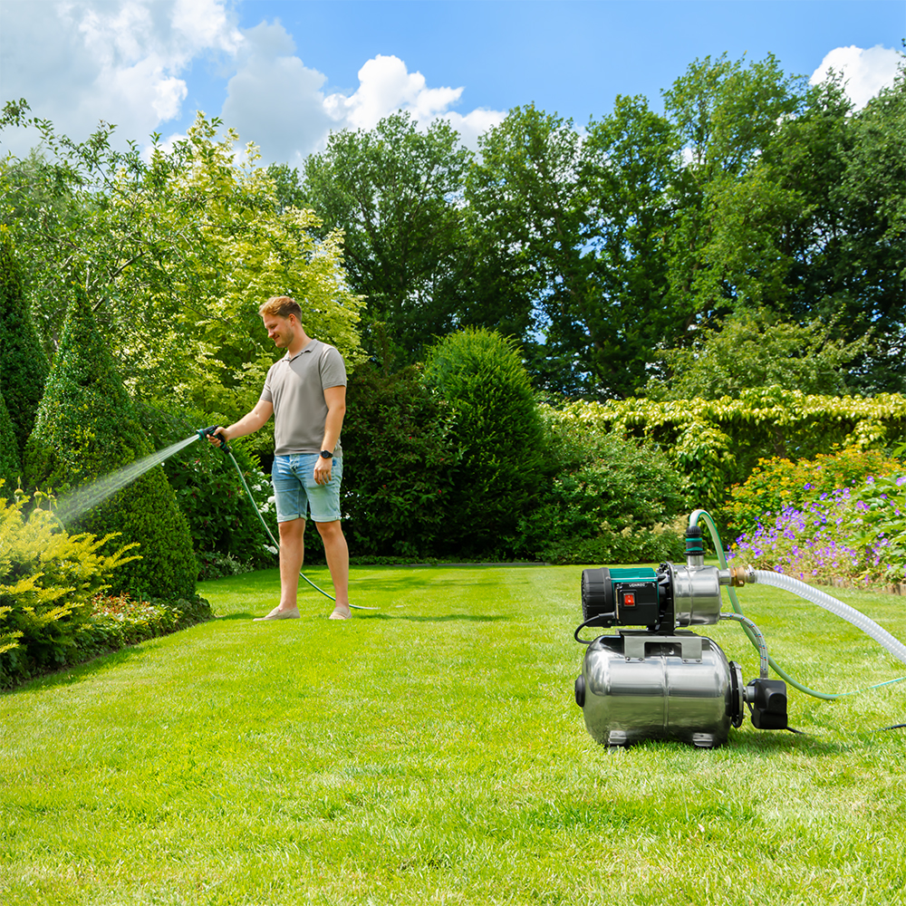 Man besproeit planten in een zonnige tuin. Groen gras, bomen en een waterpomp zijn zichtbaar. Blauwe lucht met wolken.