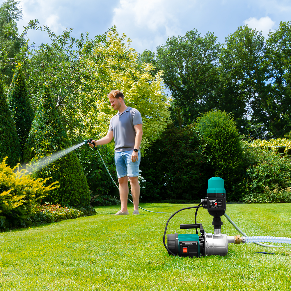 Een man besproeit een tuin met een slang. Groen gras, bomen en een waterpomp op de achtergrond. De lucht is blauw en zonnig.