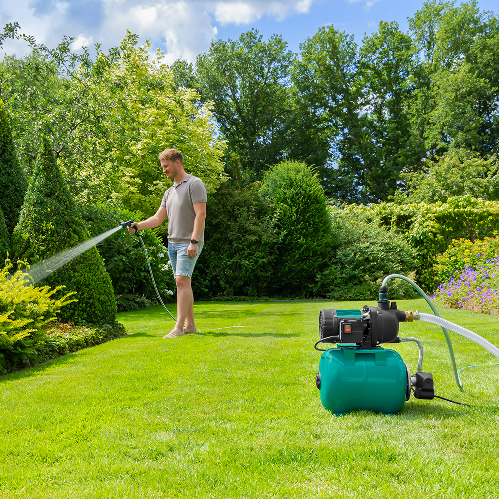 Een man besproeit een tuin met een slang. Een groene pomp staat op het gras. Bomen en struiken omringen de scène onder een blauwe lucht.
