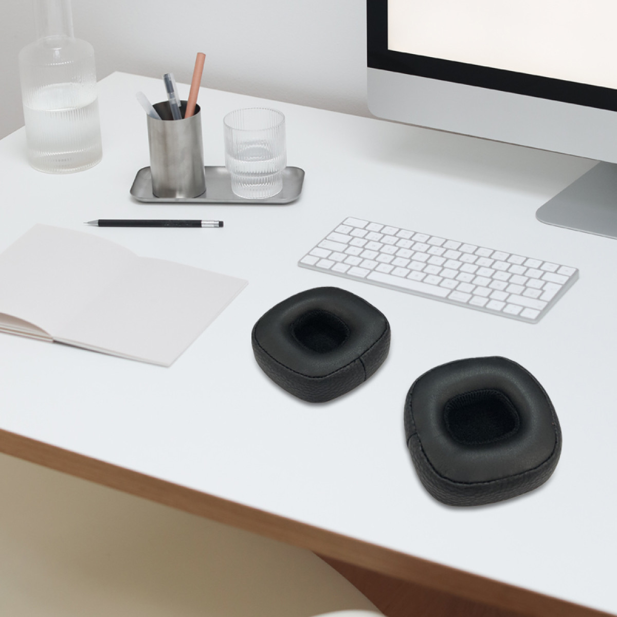 Bureau met items: monitor, toetsenbord, twee zwarte oorkussens, pennenhouder, waterfles, glas en notitieboek.