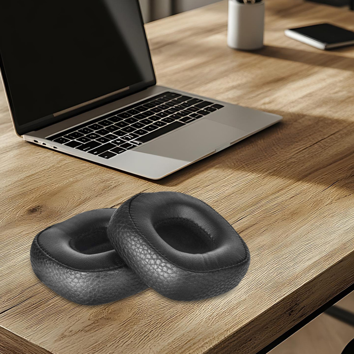Twee zwarte, ronde oorkussens liggen op een houten bureau. Een laptop op de achtergrond, met een pennenhouder en een notitieblok.