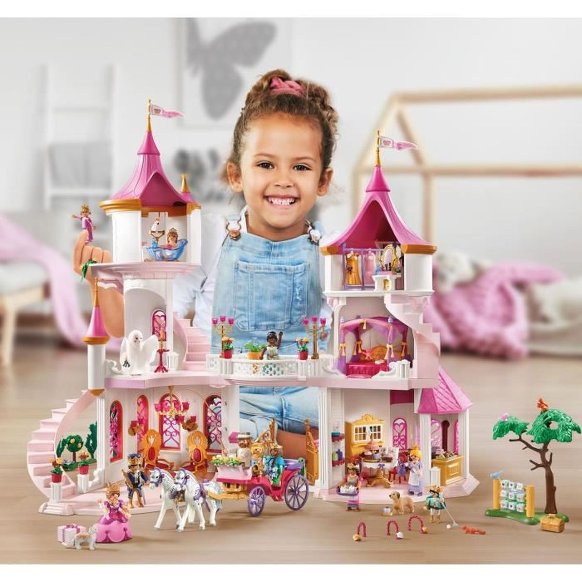 Een meisje glimlacht achter een roze kasteelspeelgoed. Het speelgoed heeft figuren, een koets en een boom. De achtergrond is een vage kamer.