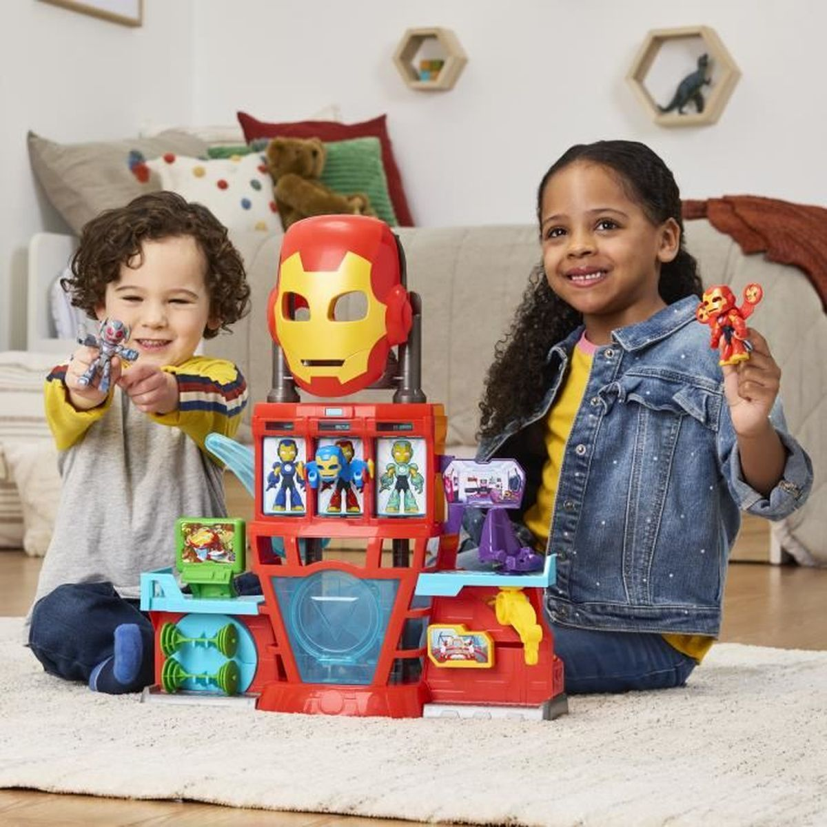Dos niños sonriendo con un juego de juguetes que presenta una máscara de Iron Man y figuras. El fondo incluye almohadas y un oso de peluche.