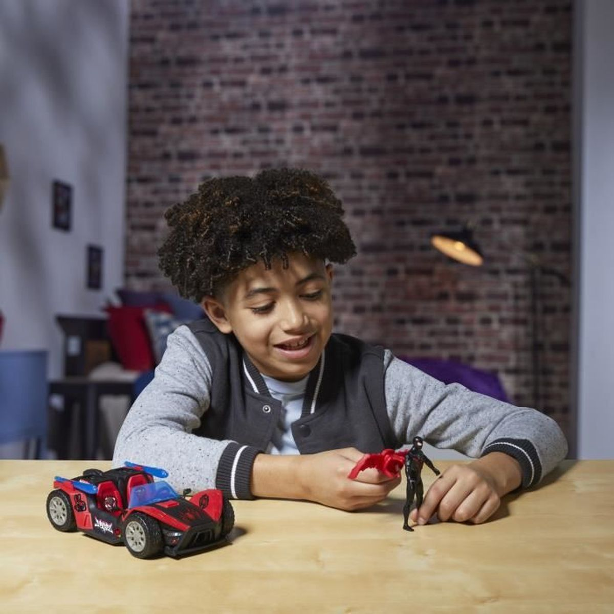 Un niño sonríe sosteniendo una figura de juguete. Un coche de juguete rojo y negro está sobre la mesa de madera. El fondo muestra una pared de ladrillos y una lámpara.