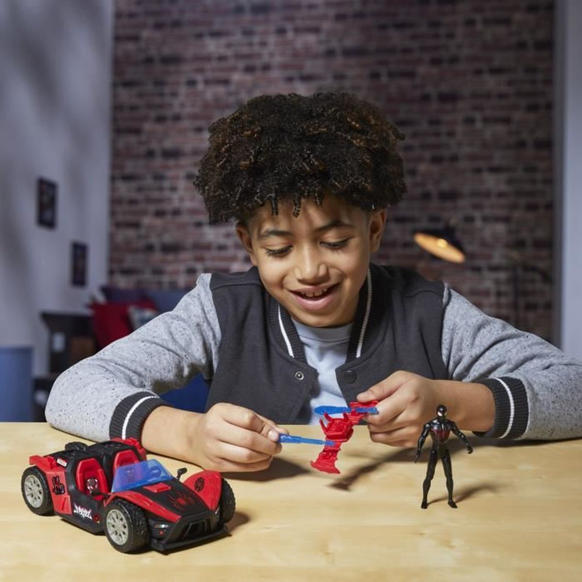 Un niño sonríe mientras juega con un coche de juguete y una figura de acción. Coche rojo y un juguete rojo y azul delante de él.