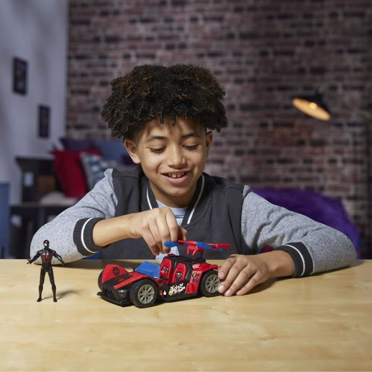 Un niño sonriente juega con un coche de juguete rojo y una pequeña figura de Spider-Man sobre una mesa de madera. El fondo muestra una pared de ladrillos.