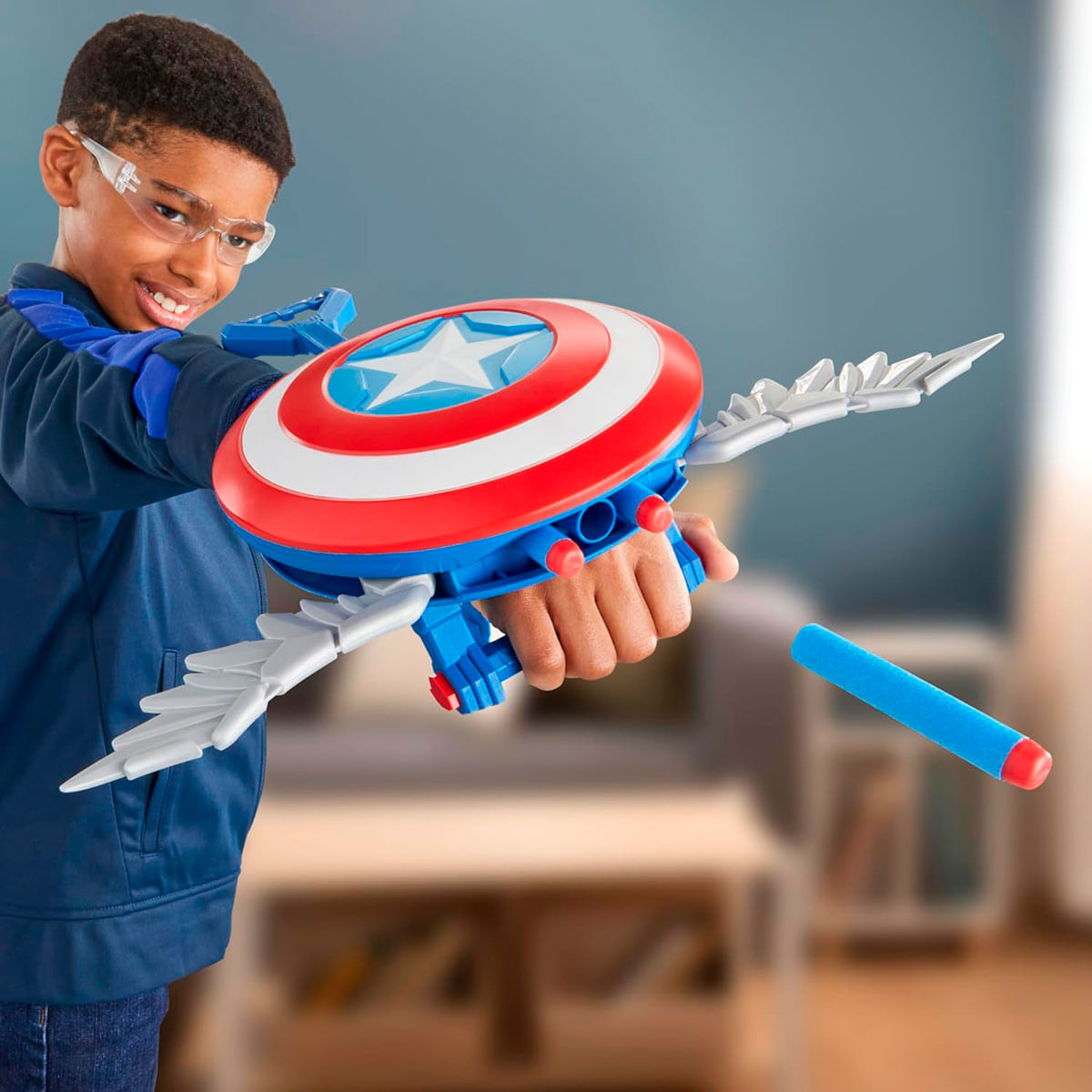 Un niño con gafas sostiene un juguete del escudo del Capitán América. El escudo es rojo, blanco y azul y tiene alas. Un dardo azul está en el aire.