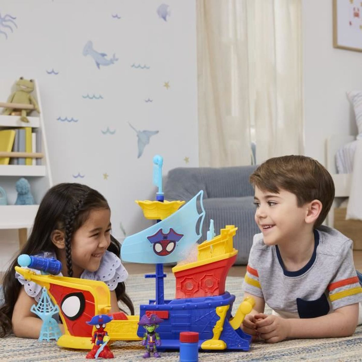Dos niños juegan con un juguete de barco pirata de Spider-Man. El barco es azul, rojo y amarillo. El fondo muestra una habitación.