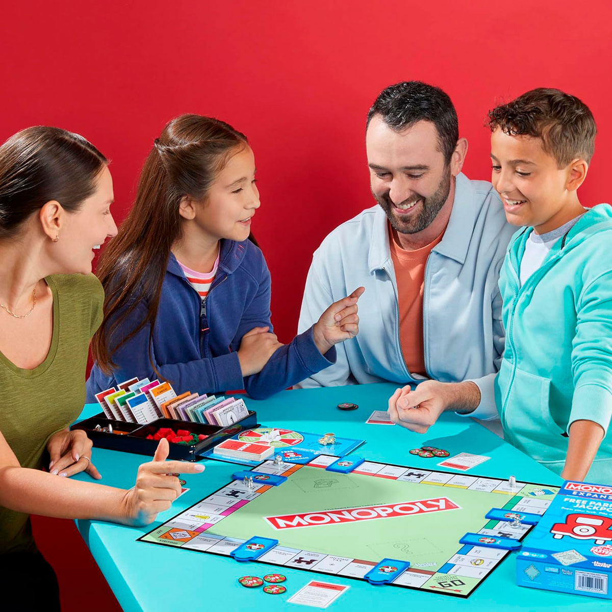 Una familia juega al Monopoly. Un tablero está sobre una mesa turquesa. Sonríen. El fondo es rojo.