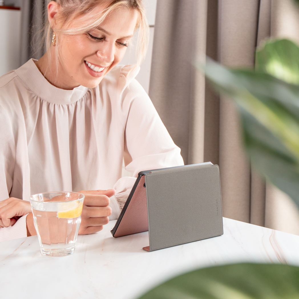 Een vrouw glimlacht terwijl ze naar een tablet kijkt. Een glas water met citroen staat op tafel. Groene plant op de achtergrond.