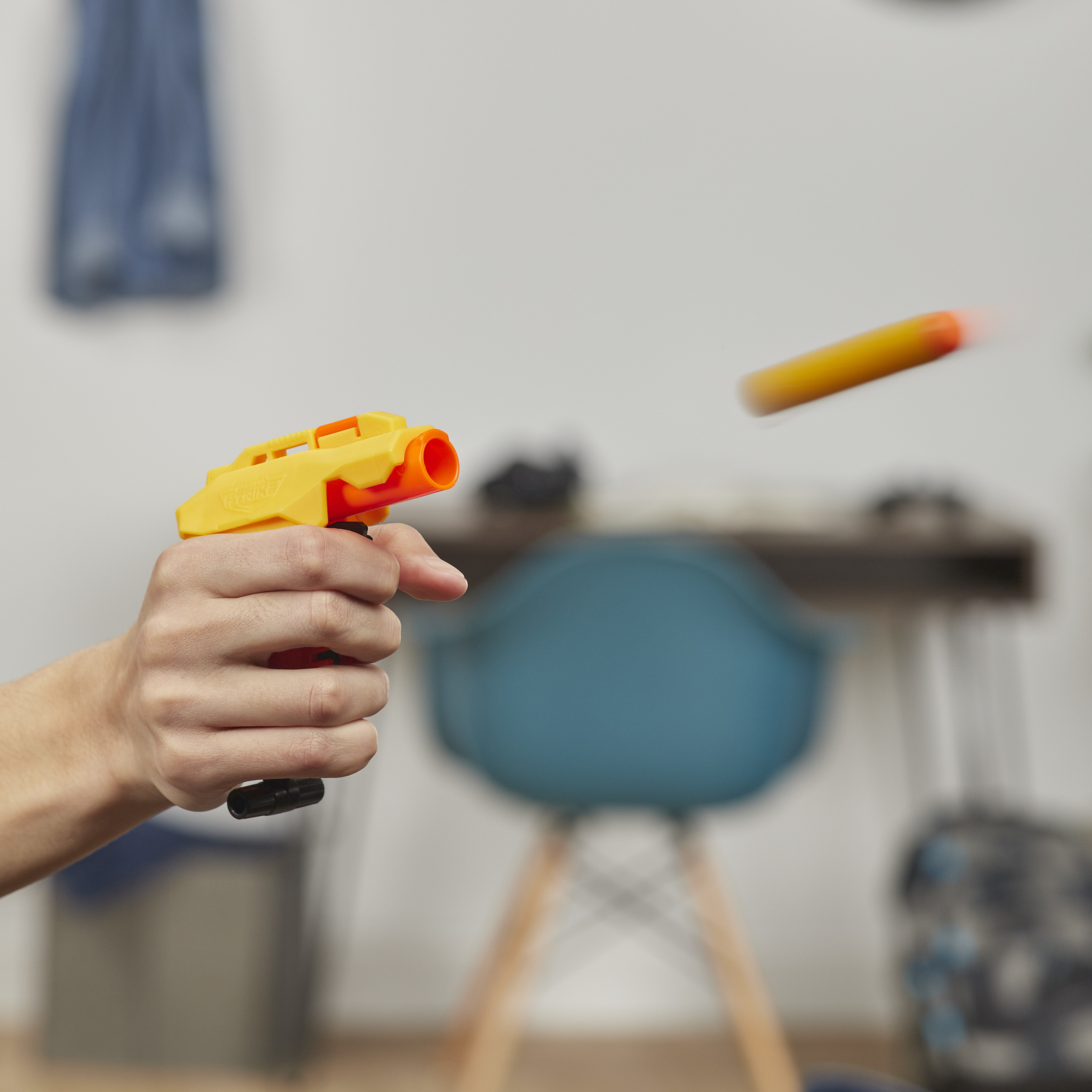 Una mano sostiene una pistola de juguete amarilla, disparando un dardo naranja. El fondo tiene una silla y un escritorio.
