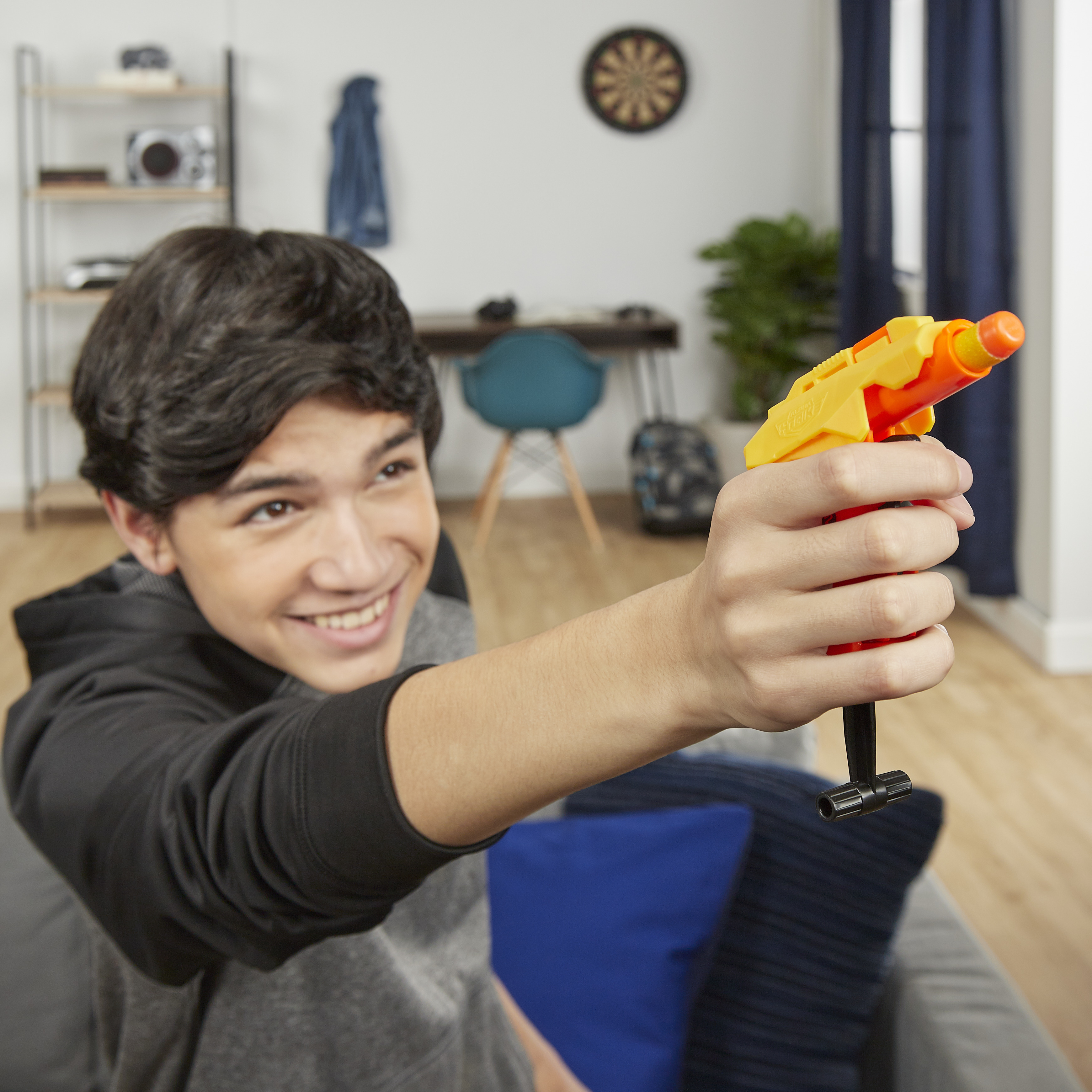 Un adolescente sonriente apunta con una pistola de juguete amarilla y naranja. El fondo muestra un salón.