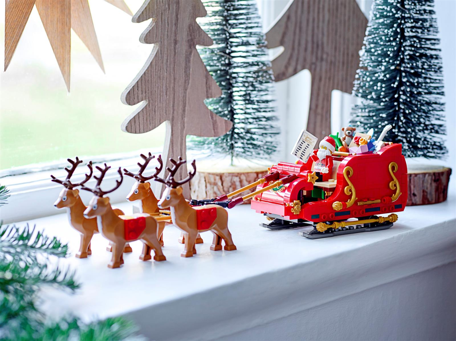 Ein roter Lego-Weihnachtsschlitten, gezogen von vier Rentieren. Der Weihnachtsmann hält eine Liste. Weihnachtsbaum und Stern im Hintergrund aus Holz.