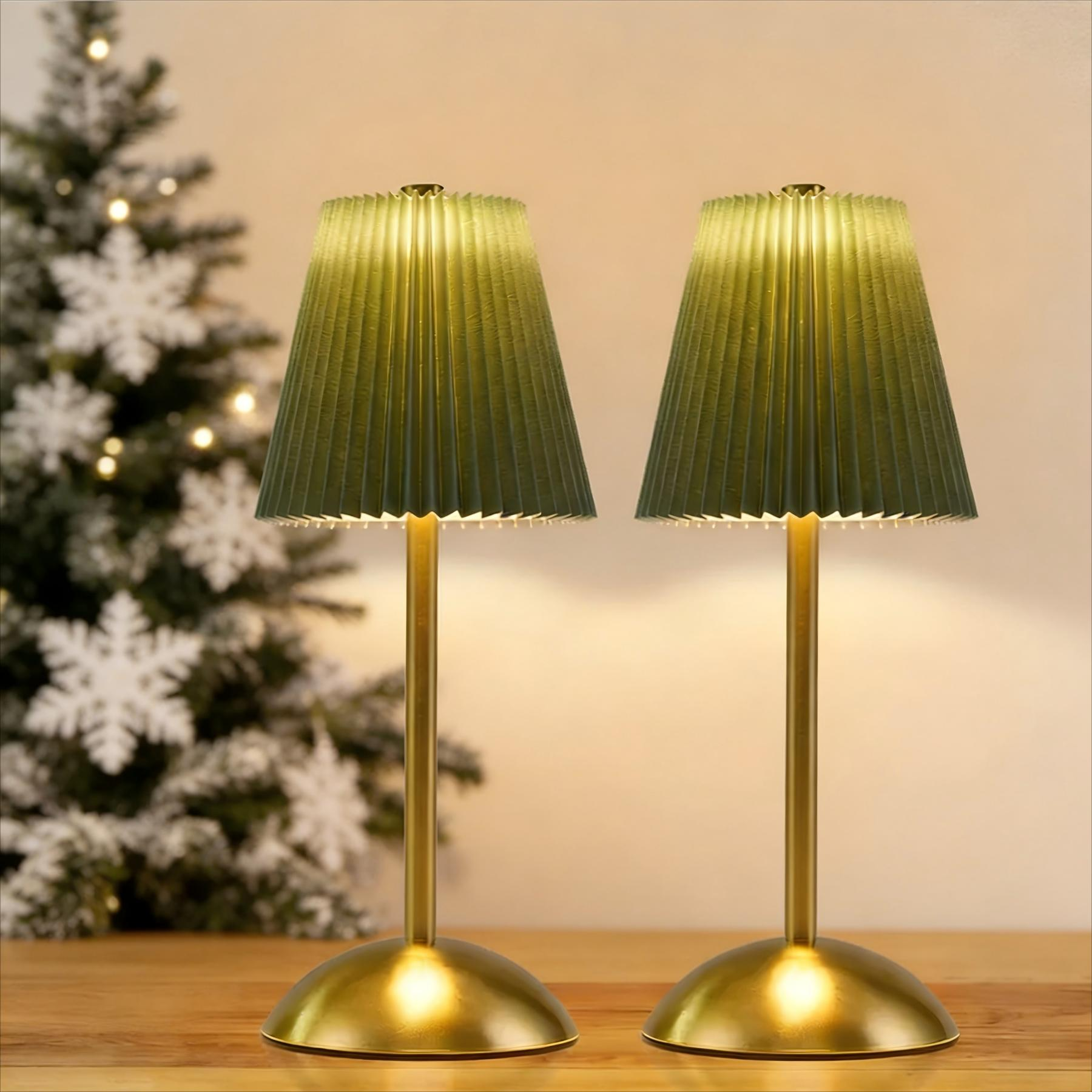 Due lampade verdi con basi dorate su una superficie in legno. Un albero di Natale illuminato sullo sfondo con fiocchi di neve.