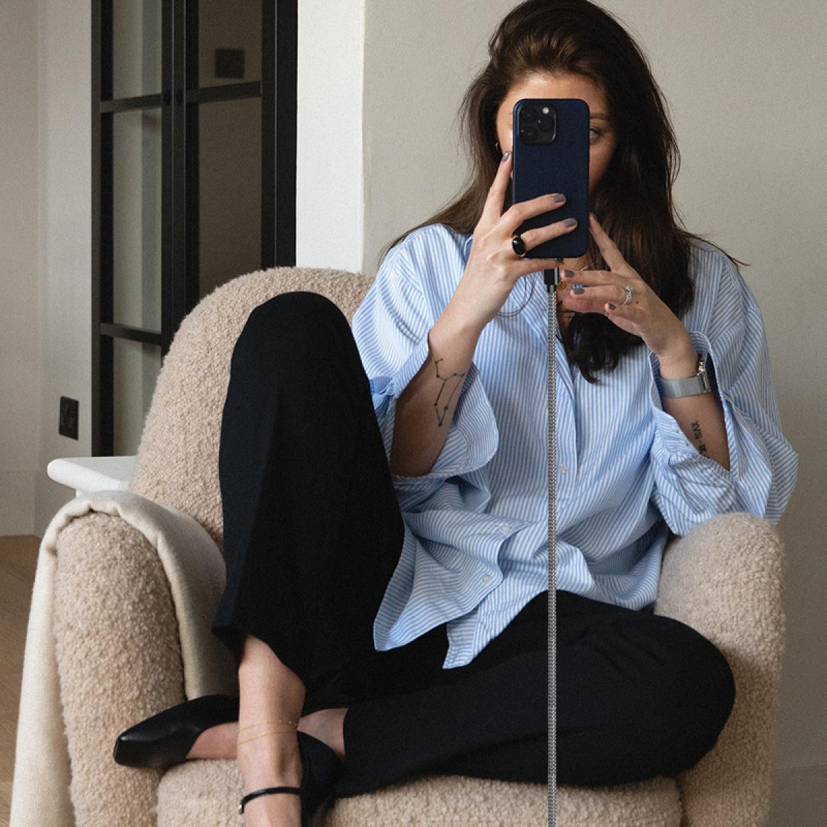 Kobieta w niebieskiej koszuli w paski i czarnych spodniach siedzi, trzymając telefon przed twarzą. Zwisa kabel.