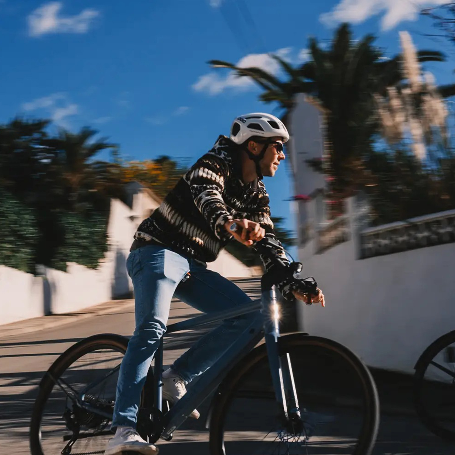 Ein Radfahrer mit Helm und Sonnenbrille fährt auf einer Straße Fahrrad. Palmen und ein blauer Himmel sind im Hintergrund.