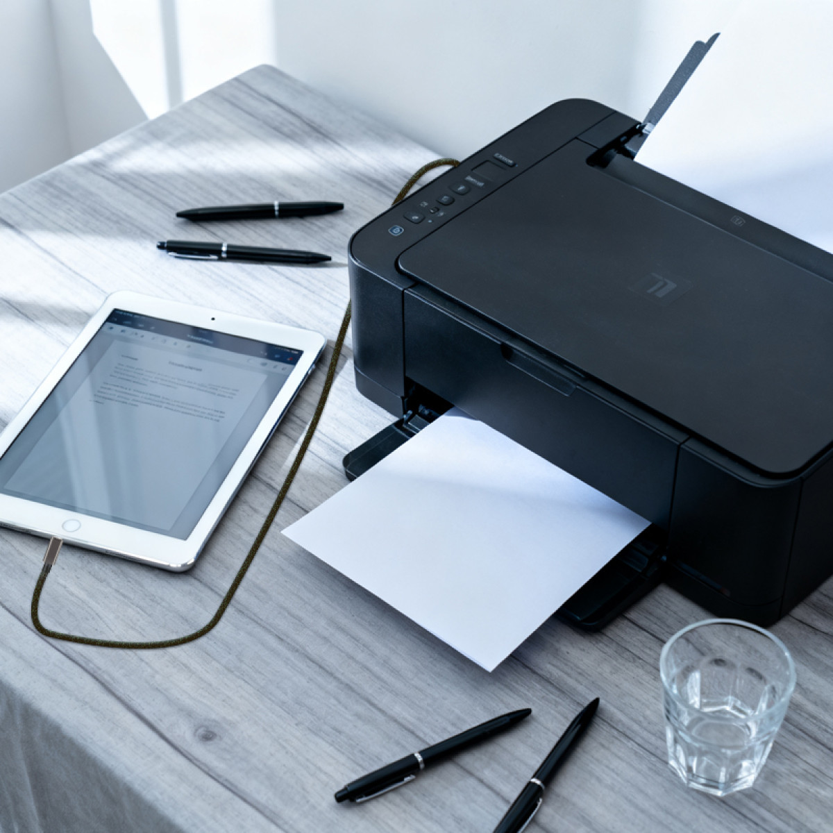 Schwarzer Drucker, der ein Papier druckt. Ein Tablet, Stifte und ein Glas Wasser liegen auf einem Holztisch.