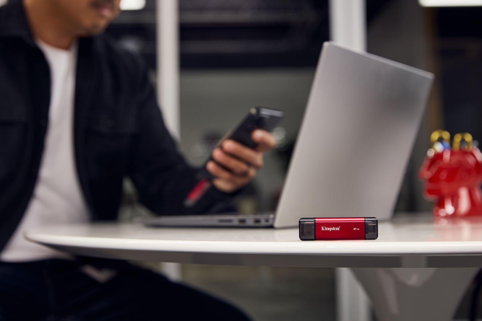 Eine Person benutzt Telefon und Laptop. Ein roter Kingston-USB-Stick liegt auf dem weißen Tisch. Ein rotes Spielzeug im Hintergrund.