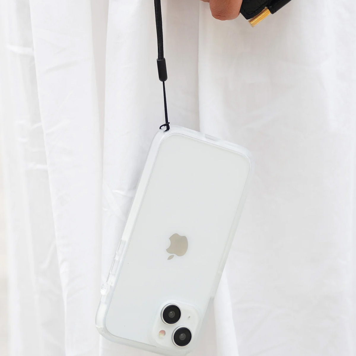 Un iPhone bianco con custodia trasparente è tenuto da un cinturino nero. Lo sfondo è tessuto bianco.