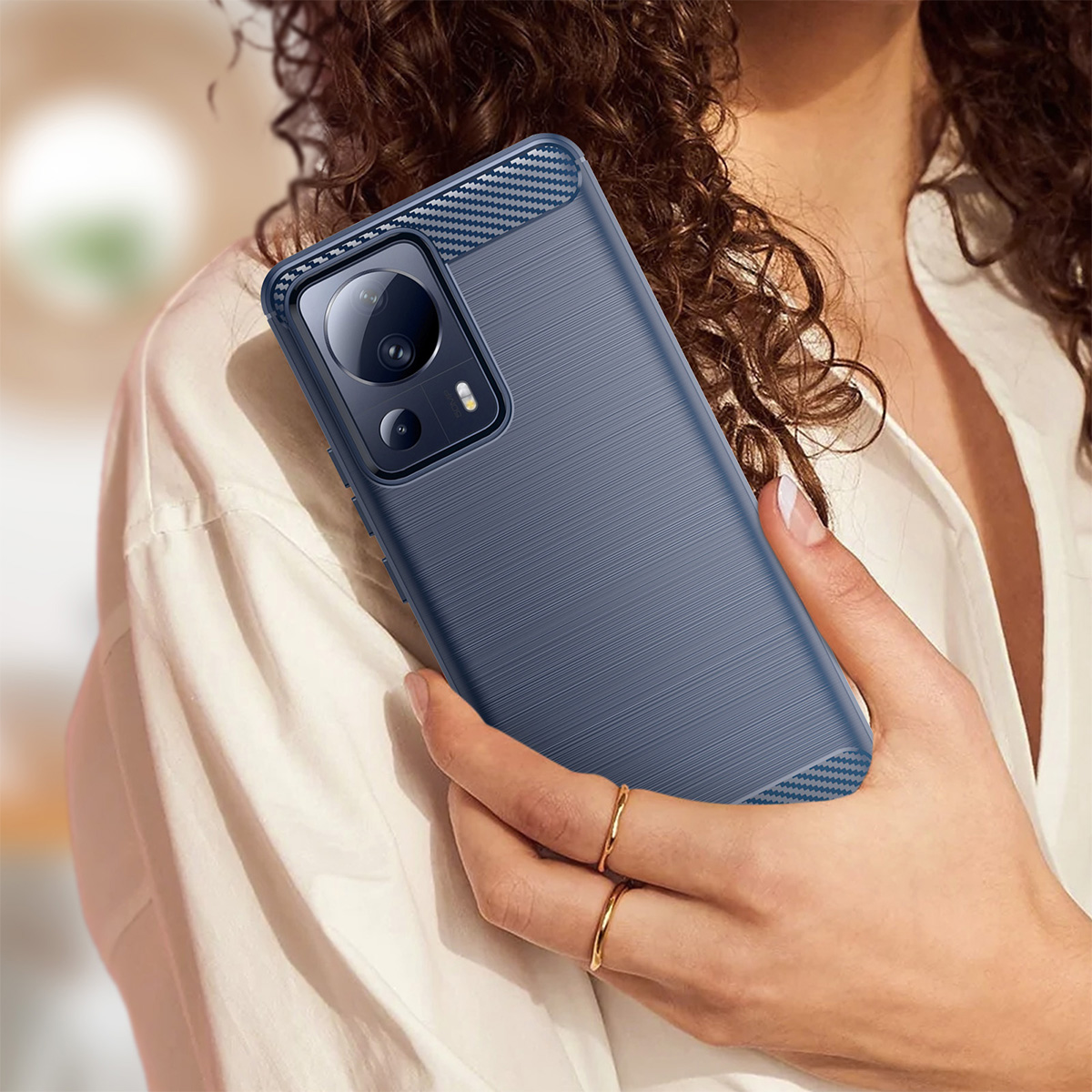 Una donna tiene in mano una custodia blu per telefono. La custodia ha una superficie strutturata e dettagli in fibra di carbonio. Indossa anelli d'oro e una camicia bianca.