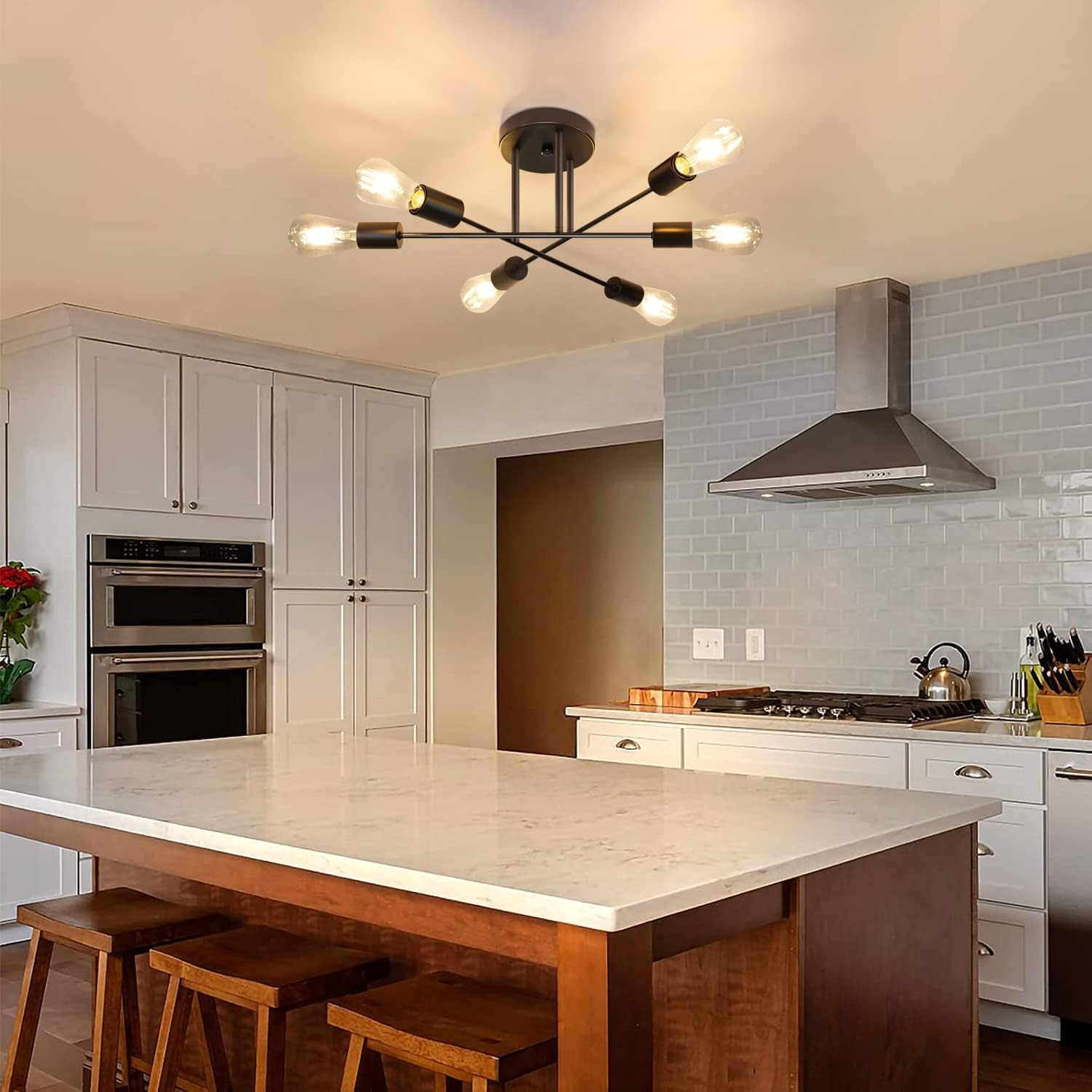 Kuchnia z centralną wyspą i stołkami. Nad głową czarne oświetlenie. Białe szafki, ceglane płytki i urządzenia ze stali nierdzewnej.