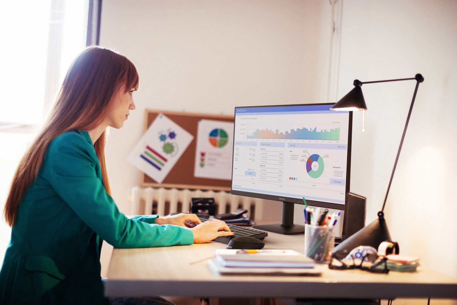 Kobieta w zielonym żakiecie pisze na klawiaturze, patrząc na ekran z wykresami. Biuro z lampą.