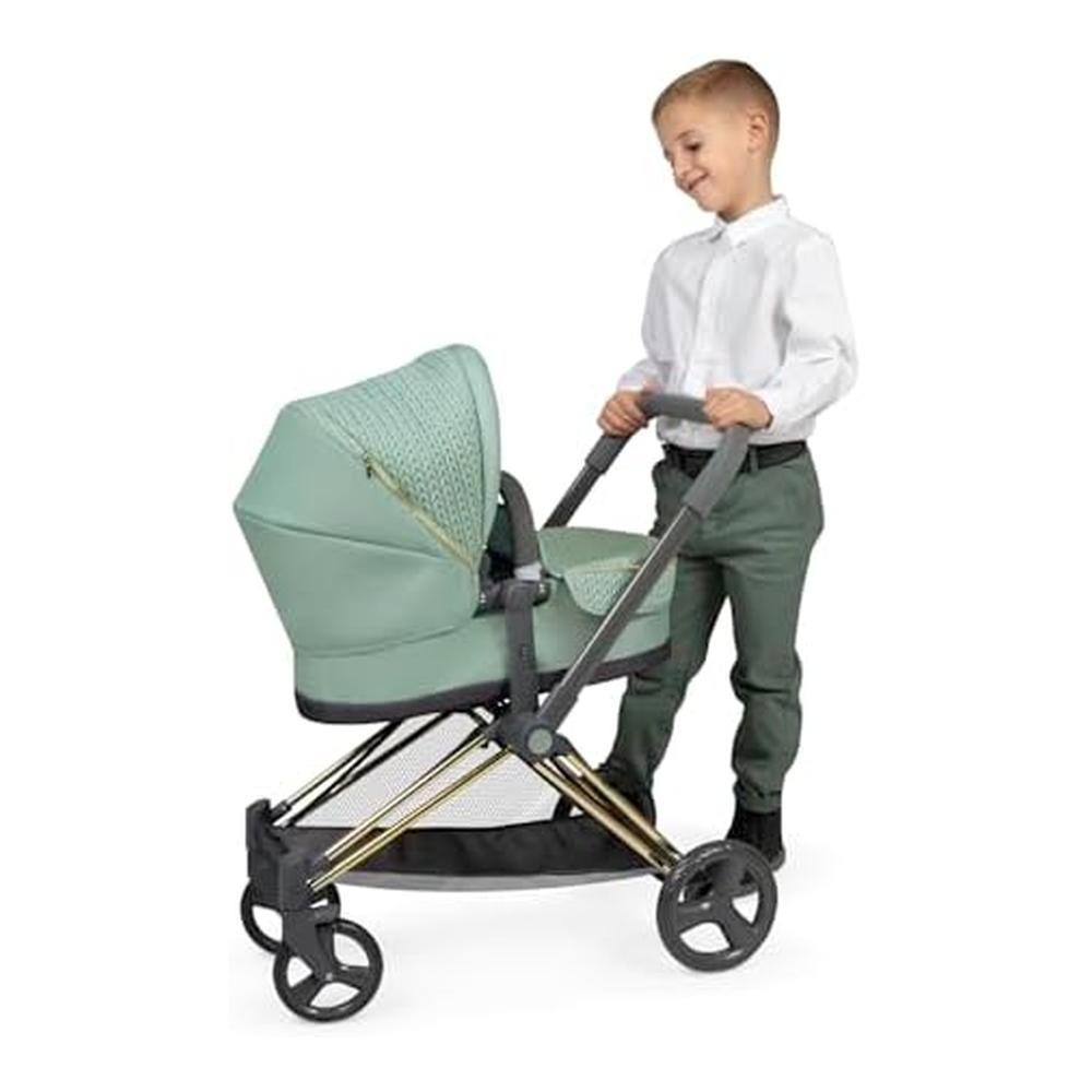 Un niño con camisa blanca y pantalón verde empuja un cochecito verde menta con detalles dorados y ruedas negras.