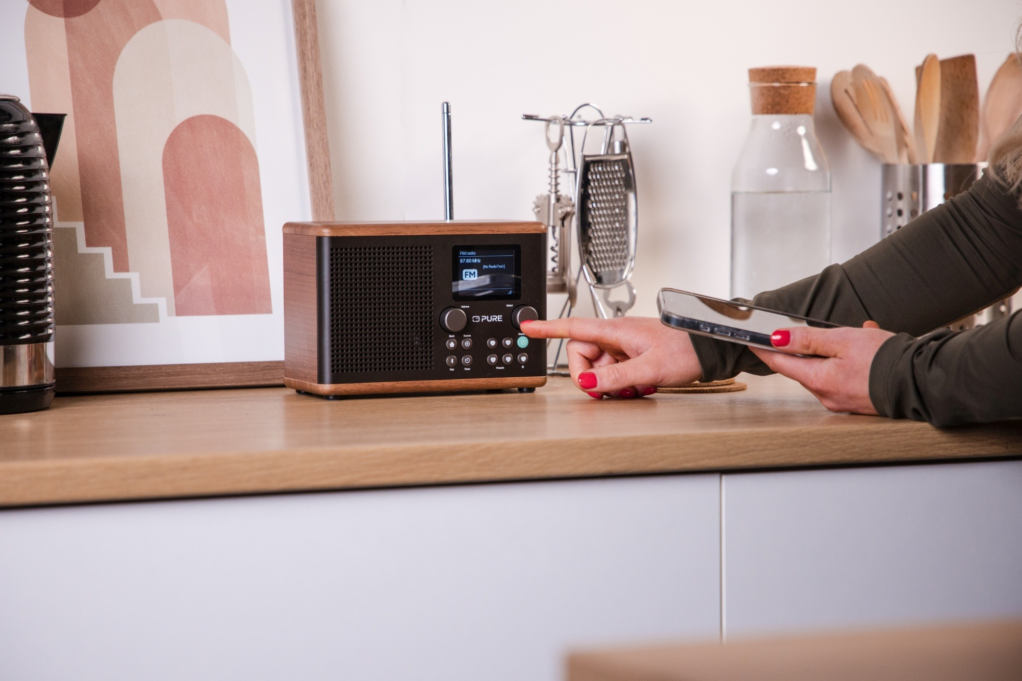 Dłoń kobiety z czerwonymi paznokciami naciska przycisk na drewnianym radiu. W drugiej dłoni telefon.