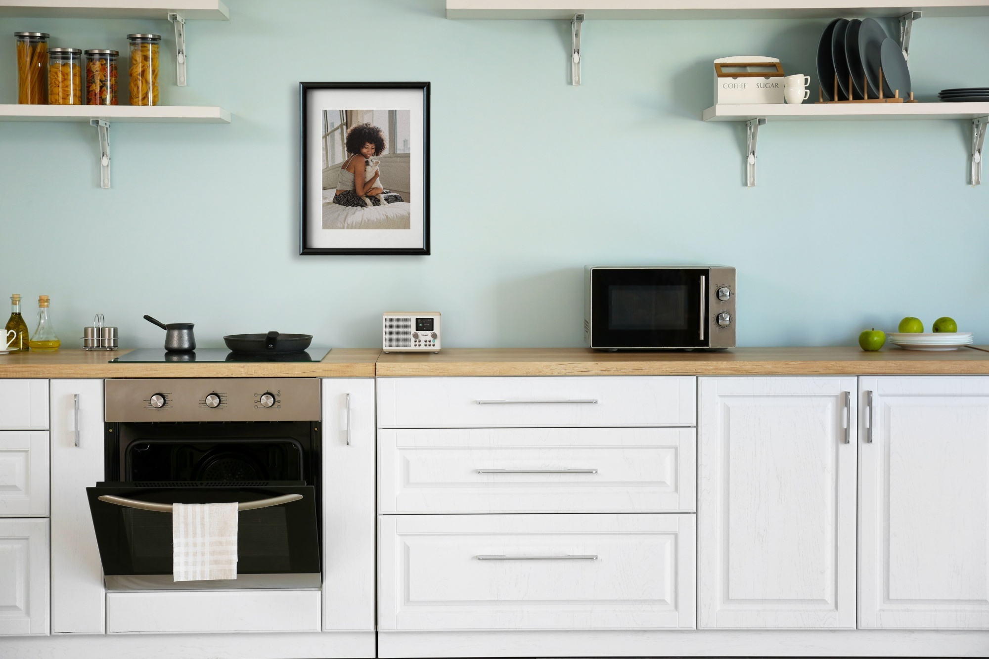Scena kuchenna z białymi szafkami, kuchenką mikrofalową i oprawionym zdjęciem. Półki z słoikami, talerzami i zestawem do kawy.