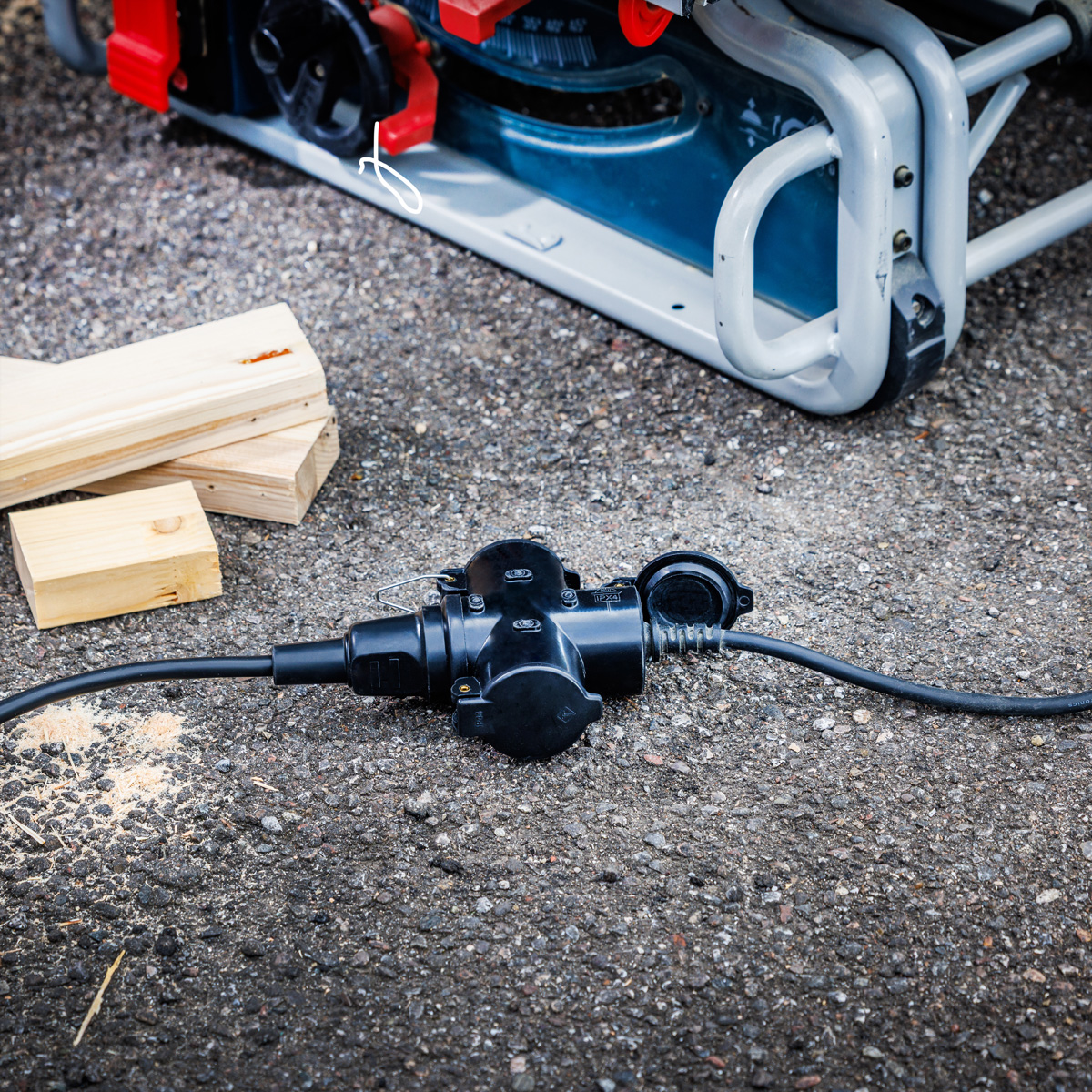 Een zwarte stroomadapter ligt op asfalt met houten blokken. Een generator is zichtbaar op de achtergrond. De scène speelt zich buiten af.