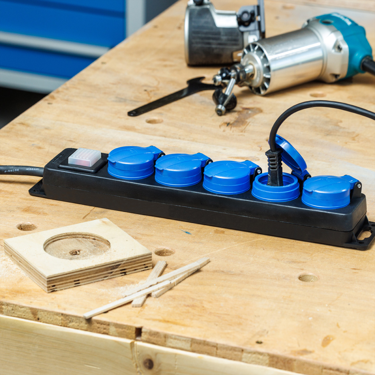 Listwa zasilająca z niebieskimi gniazdami i włożoną wtyczką. Frezarka i drewniane przedmioty na stole warsztatowym.