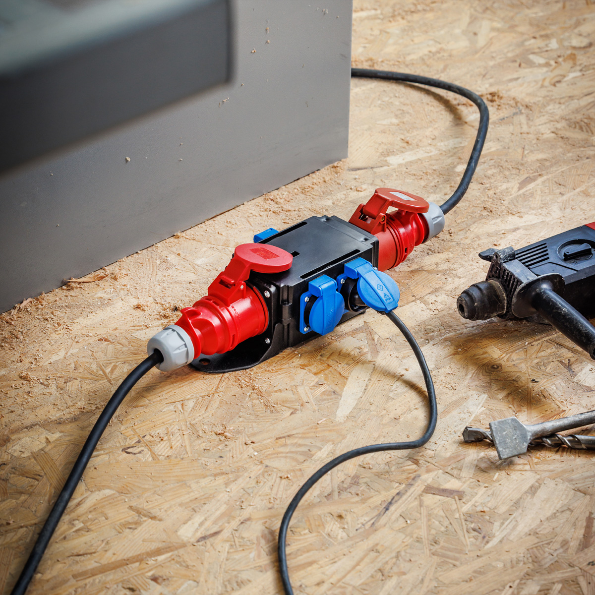 Connettori elettrici rossi e blu su legno. Un cavo nero va a una scatola grigia. Un trapano a destra.