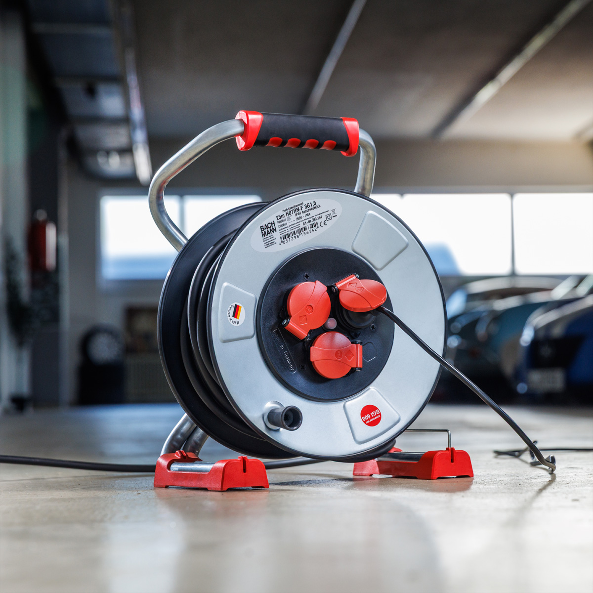 Een grijze en zwarte kabelhaspel met rode stopcontacten en handvat, in een garage. Auto's zijn zichtbaar op de achtergrond.