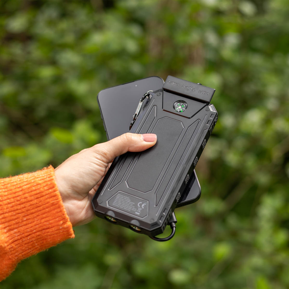 Eine Hand hält eine schwarze Powerbank mit Kompass, die ein schwarzes Telefon auflädt. Person trägt einen orangefarbenen Pullover, grüner Hintergrund.