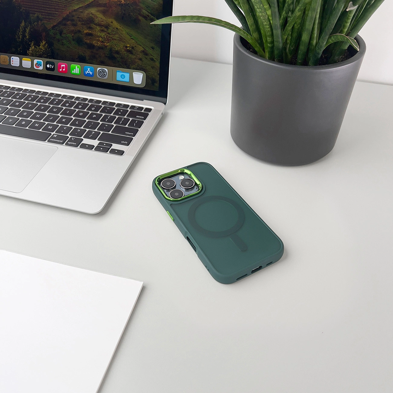 Una custodia verde, laptop e pianta su una scrivania bianca. Lo schermo del laptop mostra un paesaggio. Carta bianca.
