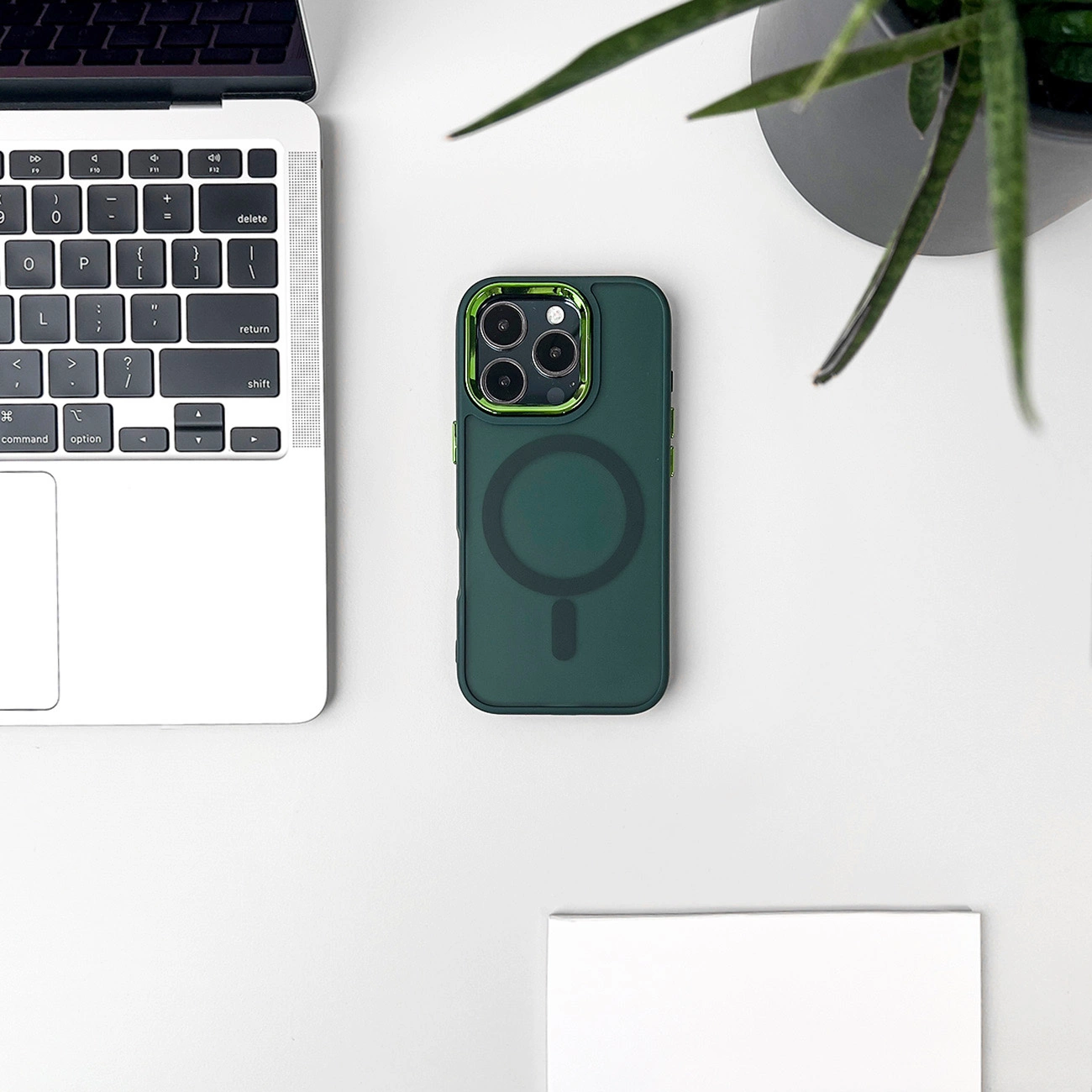 Vista dall'alto. Custodia verde per telefono con cerchio nero. Tastiera del laptop e pianta su superficie bianca.