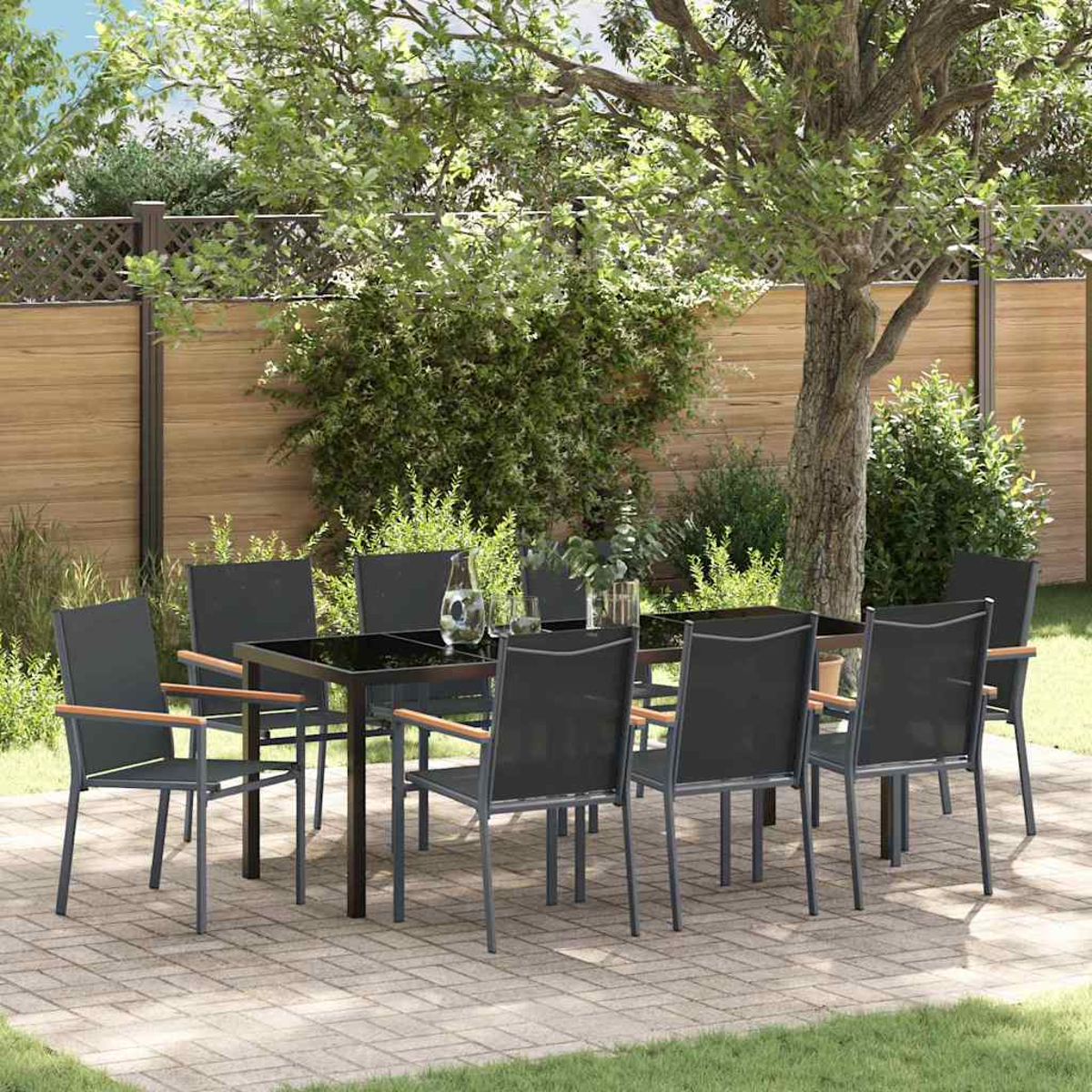 Essgruppe im Freien. Schwarzer Tisch mit acht Stühlen auf einer Terrasse. Hintergrund mit Holzzaun, Grün und Baum.