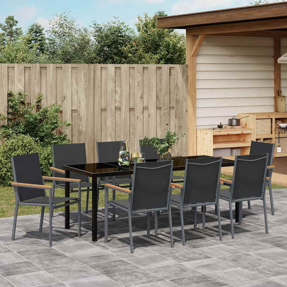 Essgruppe im Freien: schwarzer Tisch, graue Stühle mit Holzarmlehnen, auf einer gefliesten Terrasse. Hintergrund: Holzzaun und Grün.