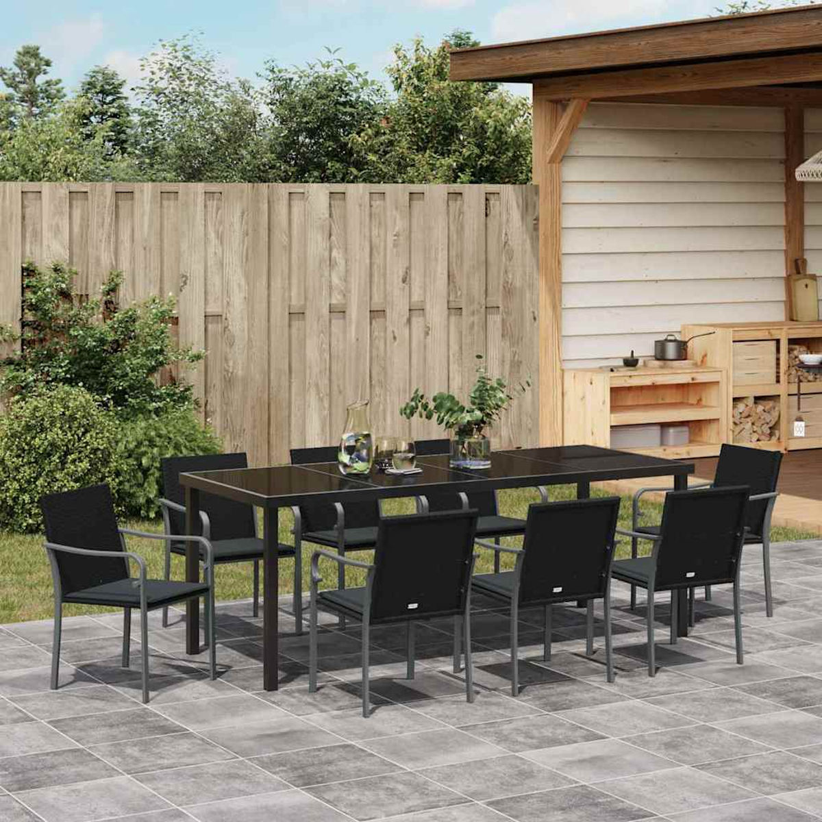 Juego de comedor al aire libre: mesa y sillas negras en un patio. Valla y estructura de madera al fondo. Plantas y cristalería en la mesa.