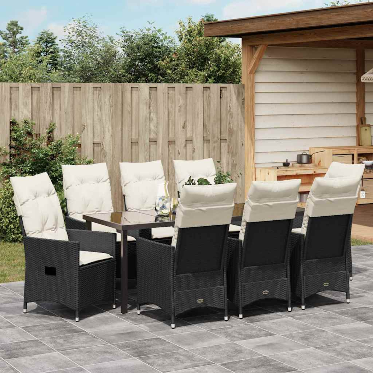 Essgruppe im Freien. Schwarze Korbstühle mit cremefarbenen Kissen um einen Tisch mit Glasplatte. Holzzaun und Struktur im Hintergrund.
