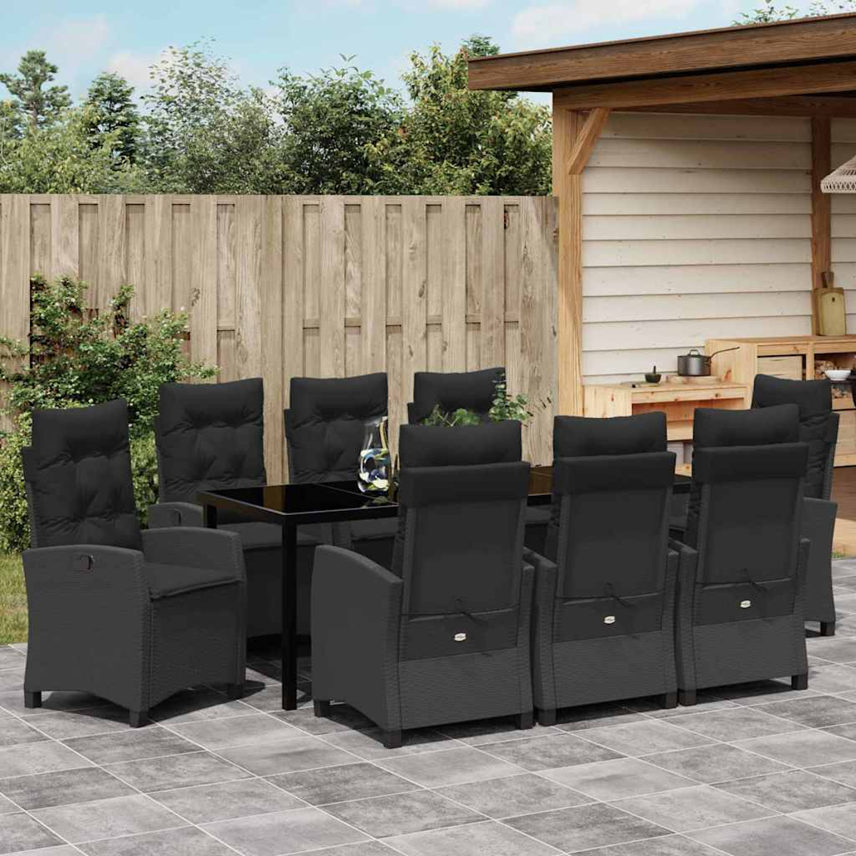 Essgruppe im Freien mit schwarzen Stühlen und einem Tisch mit Glasplatte. Auf der Terrasse mit Holzzaun und grünem Laub im Hintergrund.