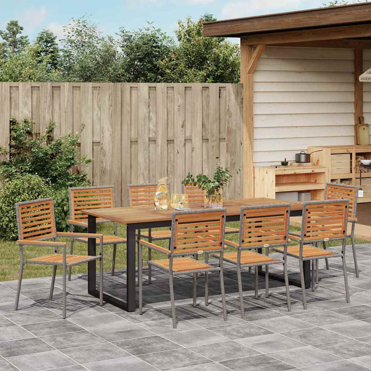 Essgruppe im Freien. Holztisch und Stühle auf einer Terrasse, gedeckt für eine Mahlzeit. Hintergrund mit Holzzaun und Bäumen.